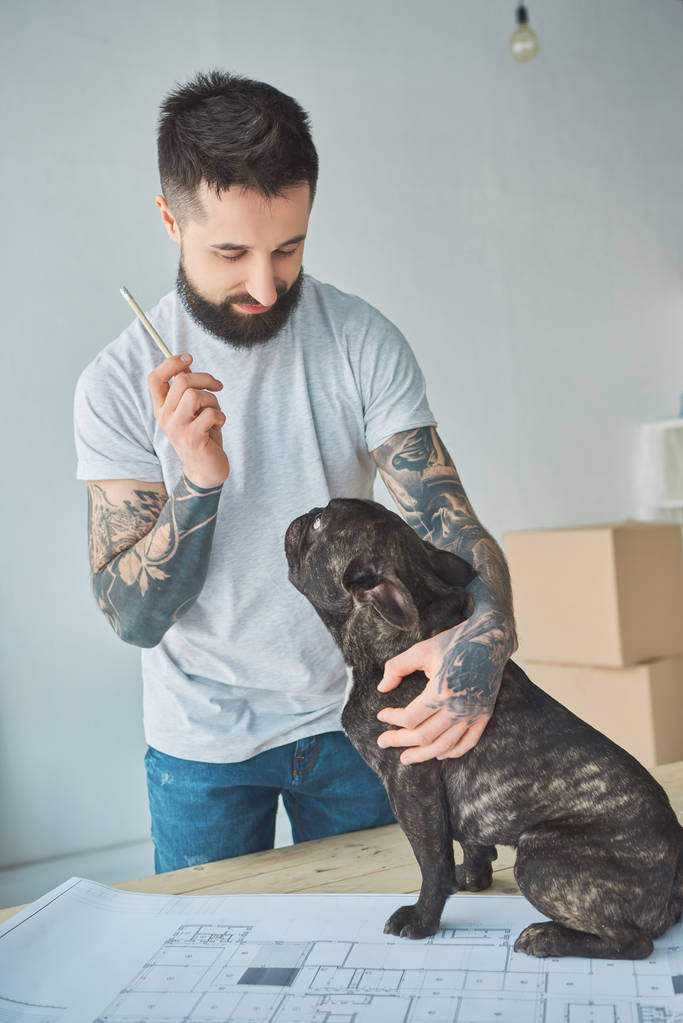Tätowierter Reparateur mit Bleistift und französischer Bulldogge steht am Entwurf der neuen Wohnung auf hölzerner Tischplatte - Foto, Bild