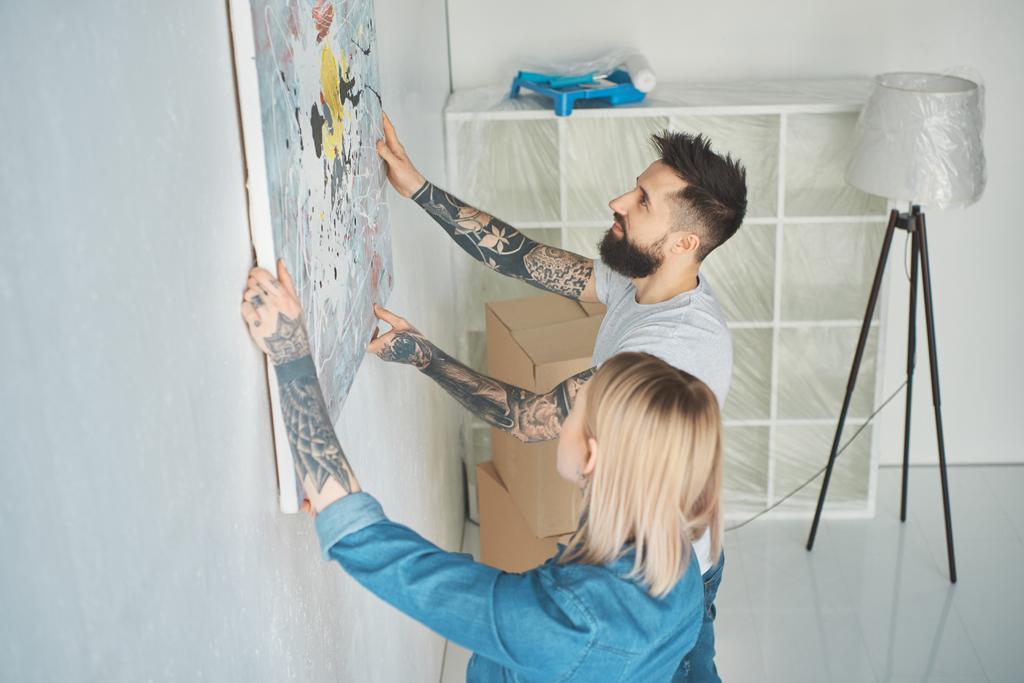 Seitenansicht eines jungen tätowierten Paares, das in seiner neuen Wohnung Gemälde an die Wand hängt - Foto, Bild