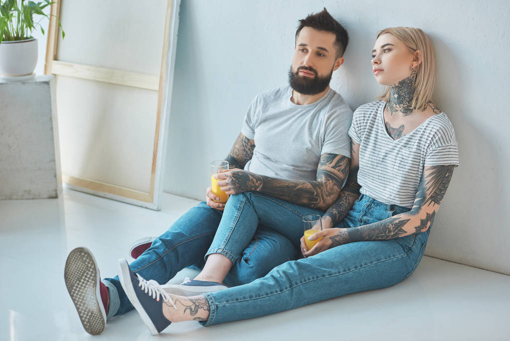 κουρασμένος τατουάζ ζευγάρι με ποτήρια χυμό κάθεται στο πάτωμα στο νέο σπίτι - Φωτογραφία, εικόνα