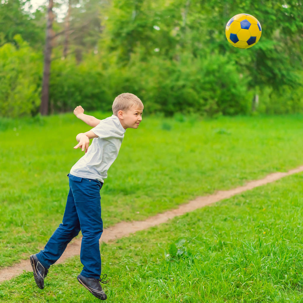 Ξανθό αγόρι σε ένα λευκό T-shirt που ρίχνουν το ποδόσφαιρο στο πάρκο - Φωτογραφία, εικόνα