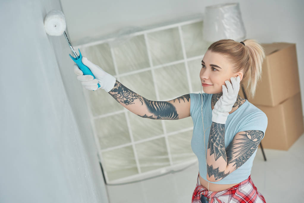 vista lateral da jovem mulher tatuada em fones de ouvido parede pintura na nova casa
 - Foto, Imagem