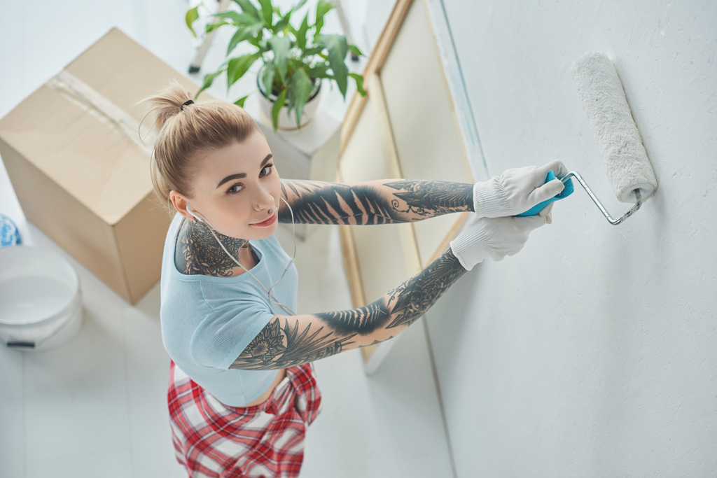 high angle view of young tattooed woman in earphones painting wall at new home - Photo, Image