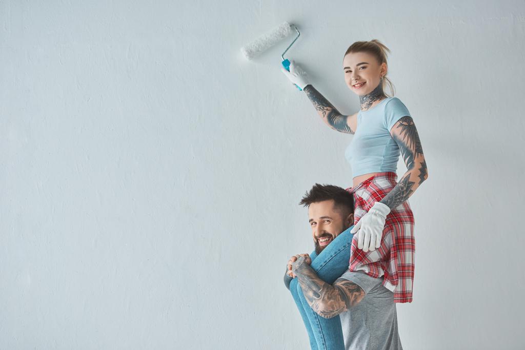 sonriente mujer con rollo pintura cepillo sentado en novios hombros mientras pintura pared en nuevo hogar
 - Foto, imagen