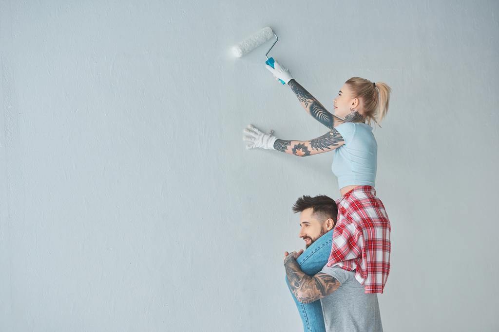 mujer con rollo pincel de pintura sentado en los hombros novios mientras que la pintura de la pared en el nuevo hogar
 - Foto, Imagen