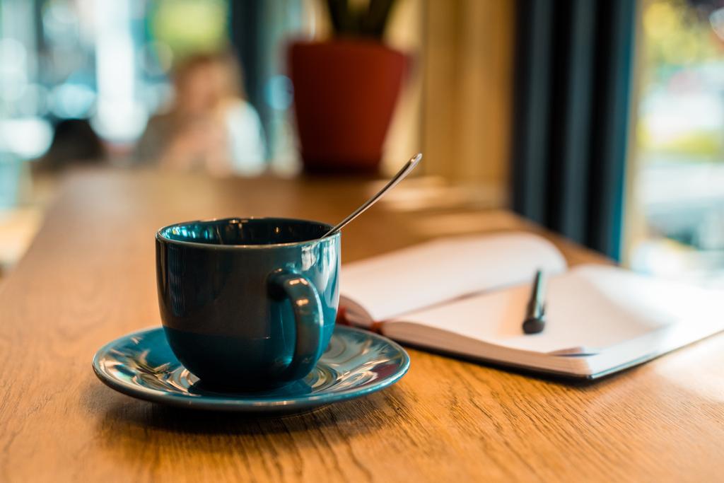kuppi kahvia lusikalla ja avaa muistikirja kynällä puinen pöytälevy kahvilassa
 - Valokuva, kuva