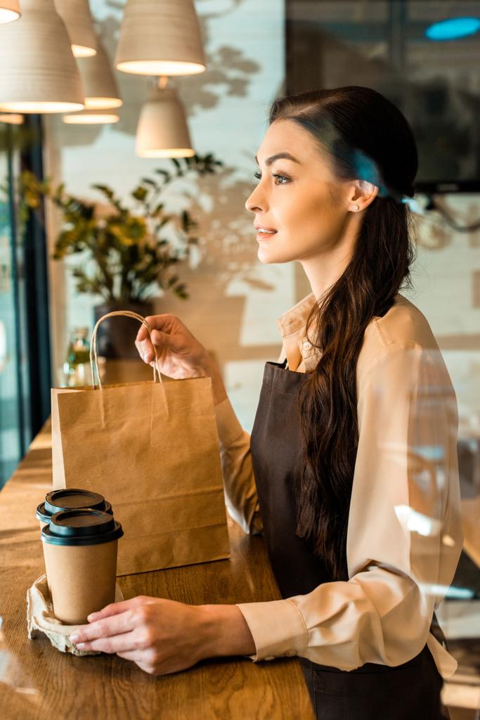 side view of beautiful waitress in apron holding disposable coffee cups and paper bag in cafe - Photo, Image