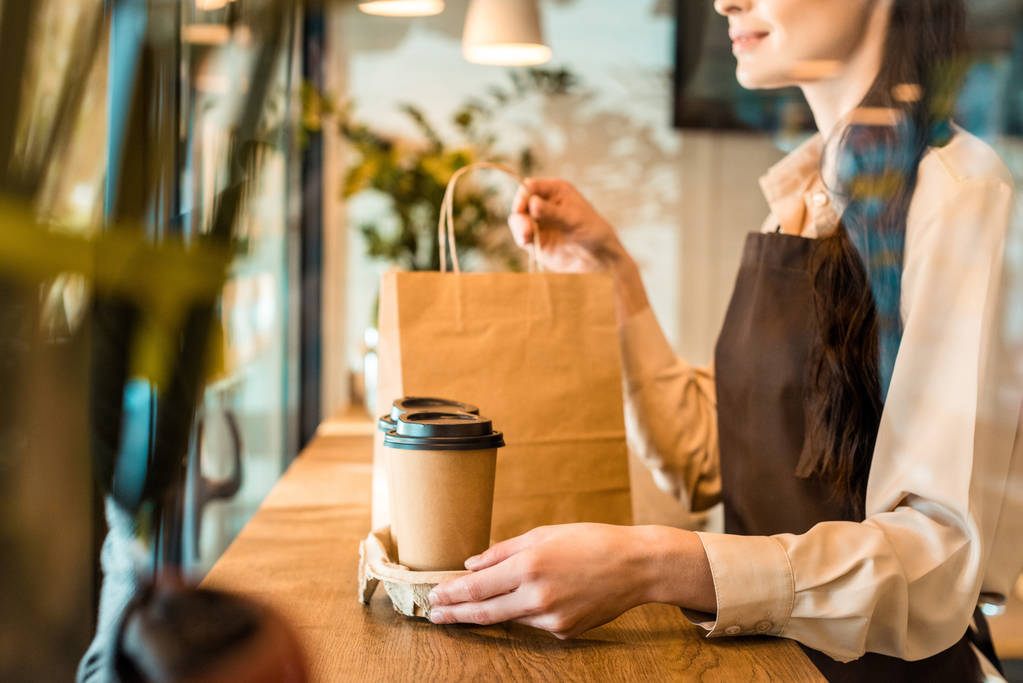 cropped image of waitress in apron holding disposable coffee cups and paper bag in cafe - Photo, Image