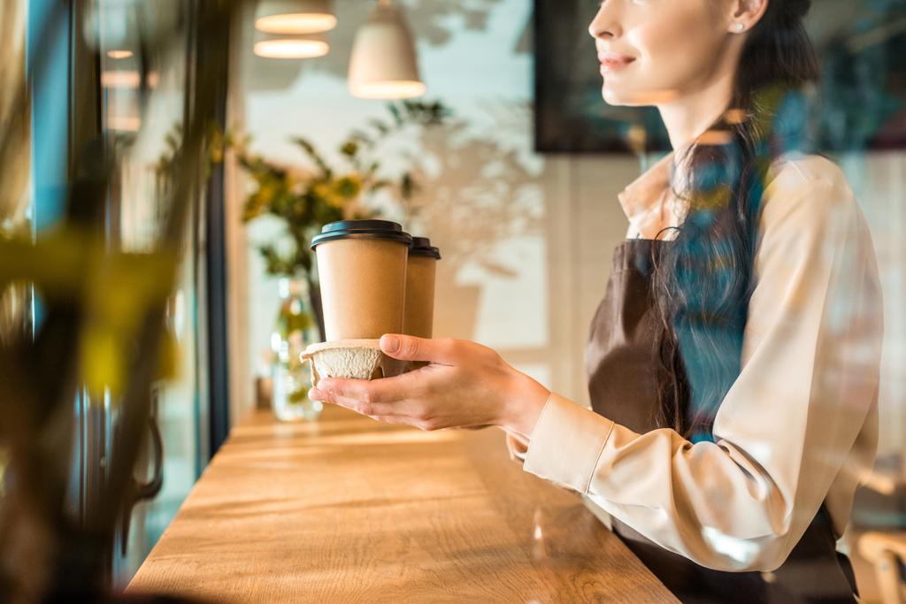 обрізане зображення офіціантки в фартусі, що тримає каву в паперових чашках в кафе
 - Фото, зображення