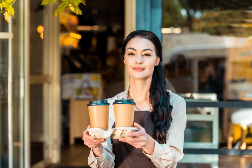 hermosa camarera en delantal sosteniendo dos café en tazas de papel cerca de la cafetería
 - Foto, imagen