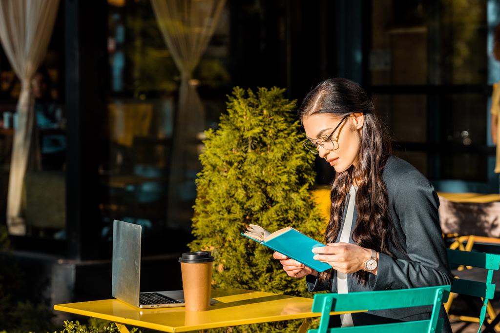 красивий фрілансер читає книгу під час перерви в ресторані тераса з ноутбуком і кавою, щоб піти
 - Фото, зображення