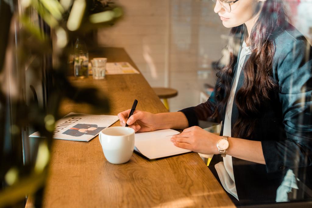 female freelancer writing in diary at table with coffee cup and business newspaper in coffee shop - Photo, Image