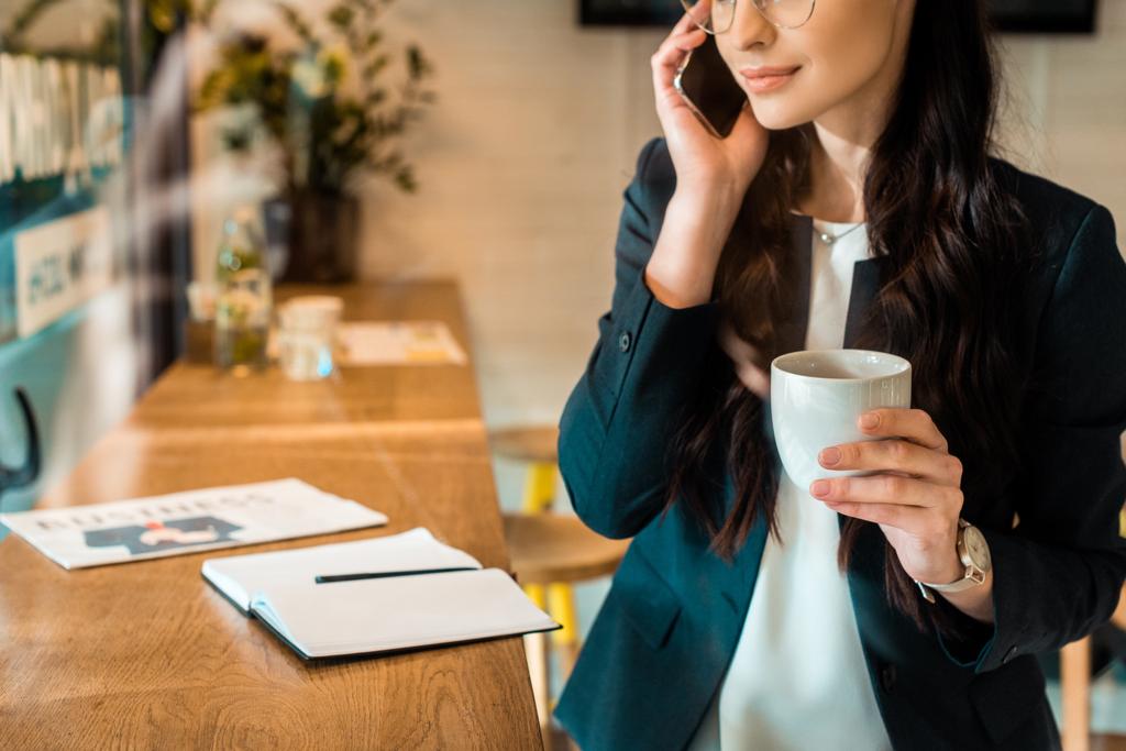 oříznutý pohled podnikatelka mluví na smartphone a drží šálek kávy v kavárně - Fotografie, Obrázek