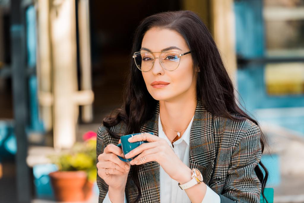όμορφη κομψή γυναίκα σε γυαλιά κρατώντας μπλε κούπα καφέ στη βεράντα του εστιατορίου - Φωτογραφία, εικόνα