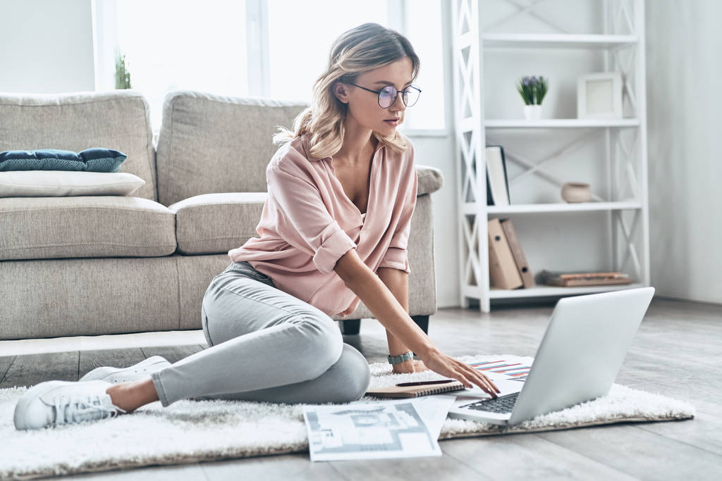 Jeune femme réfléchie dans les lunettes de travail et l'utilisation de l'ordinateur tout en revêtements de sol à la maison
           - Photo, image