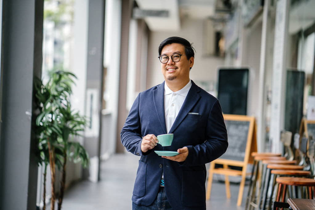Retrato de un hombre asiático chino educado y de mediana edad en espectáculos casuales y de negocios. Él está sosteniendo y bebiendo té de una elegante taza y platillo durante el día en la ciudad
. - Foto, Imagen