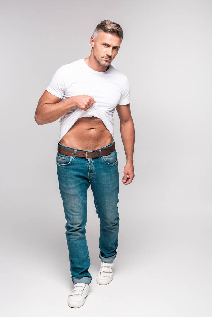 Вид в полный рост красивого мужчины в белой футболке и джинсовых штанах, смотрящего в камеру на сером
 - Фото, изображение