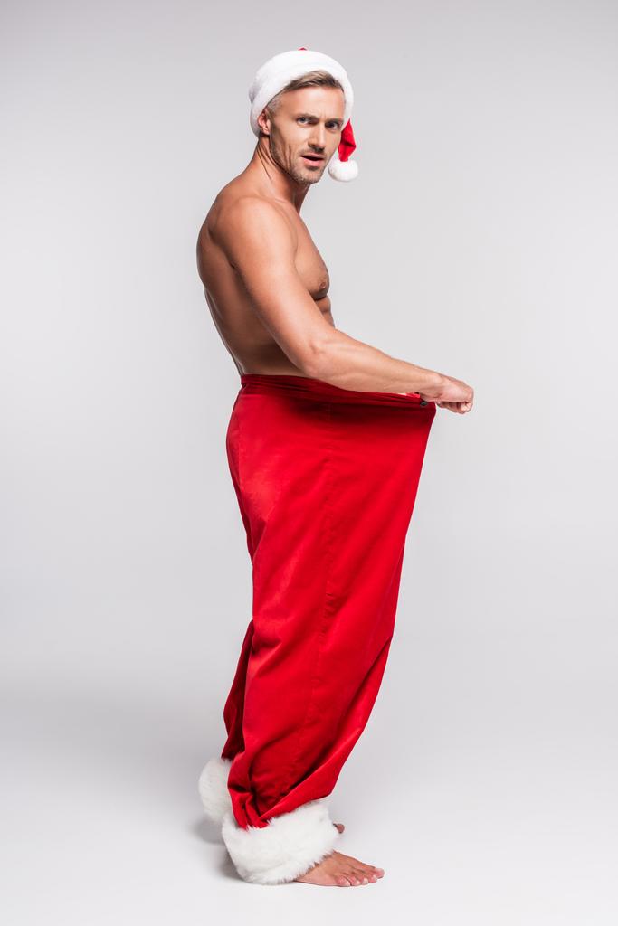 vue latérale de l'homme torse nu en santa chapeau tenant pantalon rouge et regardant la caméra sur gris
  - Photo, image