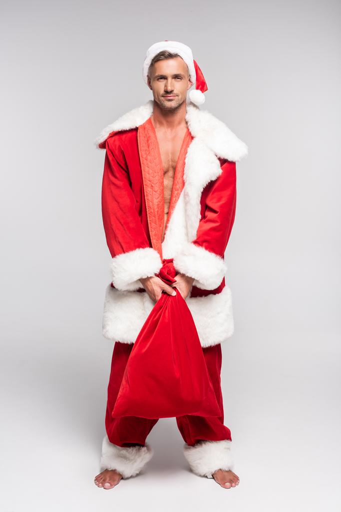 visão de comprimento total de homem descalço bonito em Santa traje segurando saco vermelho e sorrindo para a câmera em cinza
 - Foto, Imagem