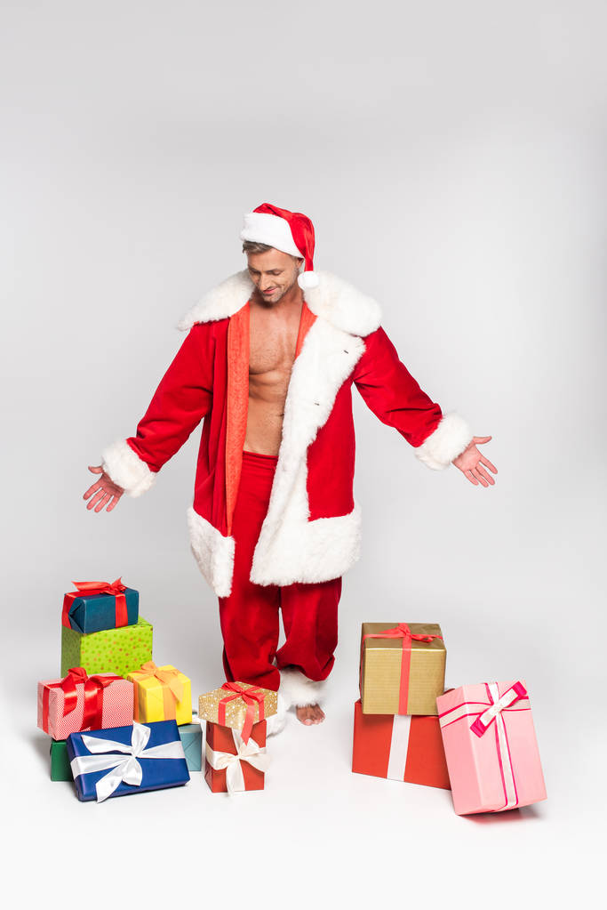 schöner Mann im Weihnachtsmannkostüm schaut auf Geschenkschachteln auf grau - Foto, Bild