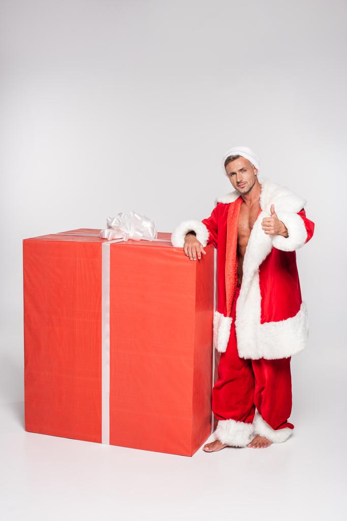 Красивый Санта Клаус, опирающийся на большую подарочную коробку и показывающий большой палец на сером
 - Фото, изображение