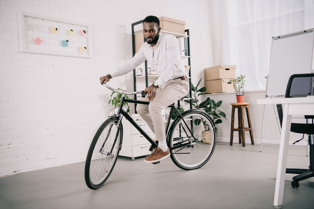 schöner afrikanisch-amerikanischer Geschäftsmann im weißen Hemd, der im Büro Fahrrad fährt - Foto, Bild
