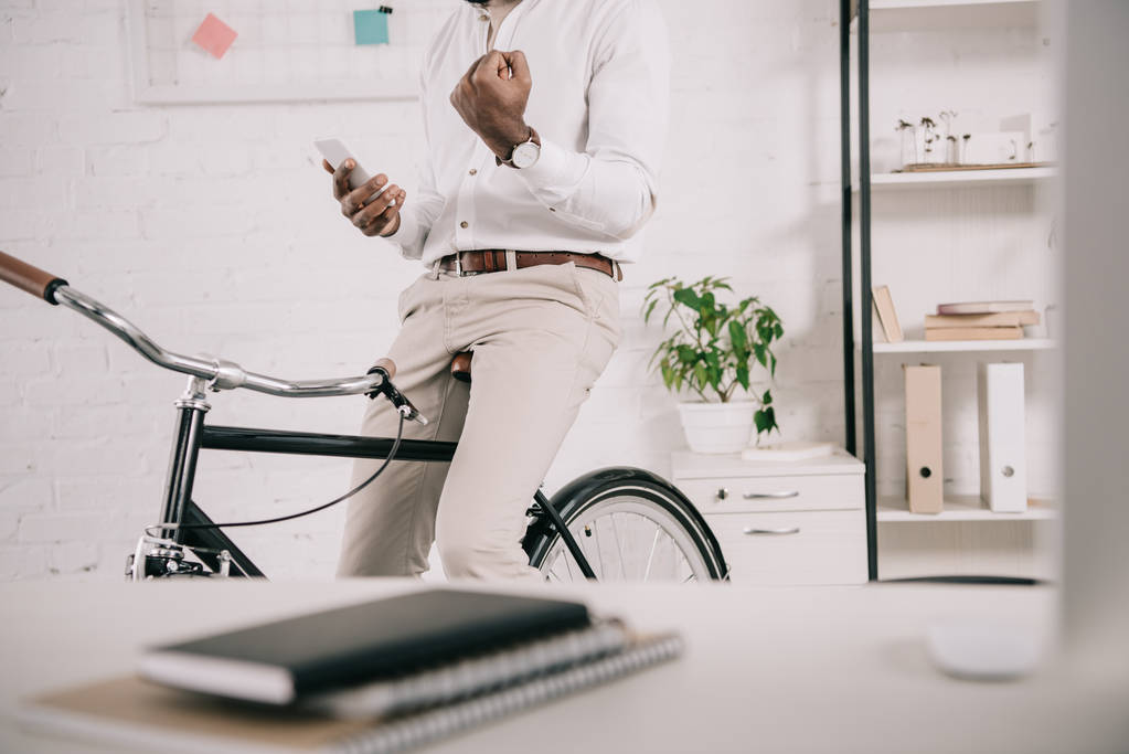 cropped image of african american businessman sitting on bike, using smartphone and showing yes gesture in office - Photo, Image