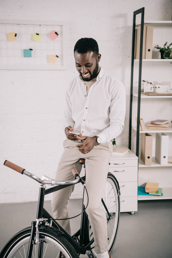 lachende knappe Afro-Amerikaanse zakenman op fiets zit en smartphone gebruiken in office - Foto, afbeelding