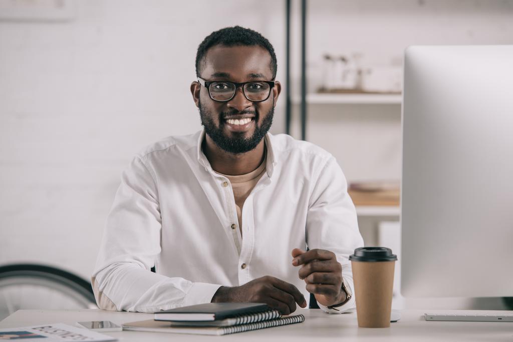 lachende knappe Afro-Amerikaanse zakenman in glazen aan tafel zitten en kijken naar camera in kantoor - Foto, afbeelding