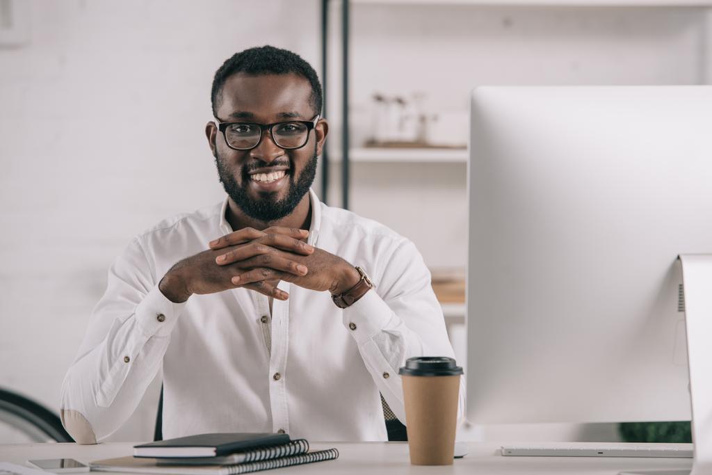 усміхнений красивий афроамериканський бізнесмен сидить за столом і дивиться на камеру в офісі
 - Фото, зображення