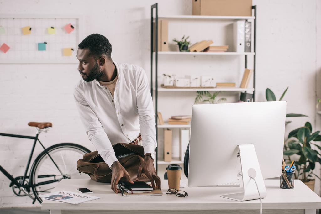 красивый африканский американский бизнесмен берет блокноты со стола и смотрит в сторону в офисе
 - Фото, изображение