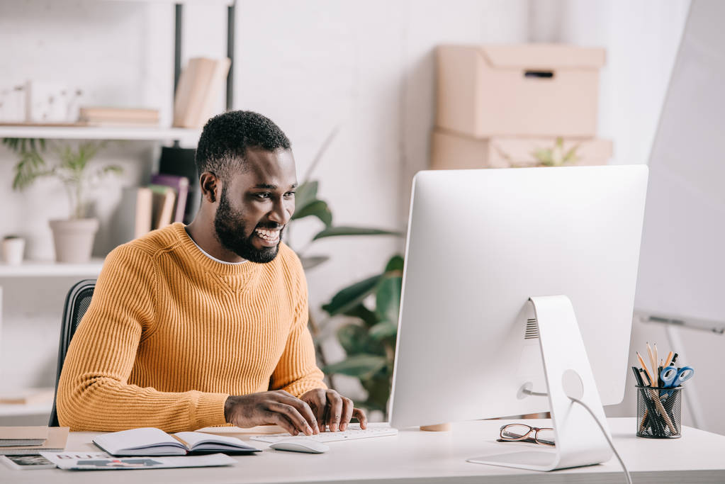 glimlachend knappe Afrikaanse Amerikaanse ontwerper in oranje trui werken op de computer in kantoor - Foto, afbeelding