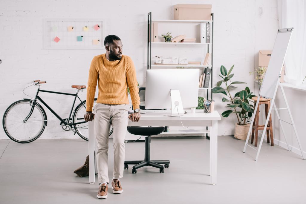 komea afrikkalainen amerikkalainen suunnittelija oranssissa puserossa nojaa pöydälle ja katsoo pois toimistossa
 - Valokuva, kuva