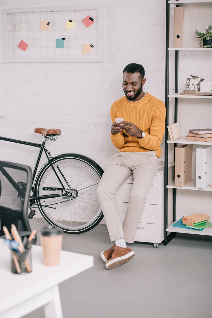 sonriente diseñador afroamericano en suéter amarillo usando teléfono inteligente en la oficina con bicicleta
 - Foto, Imagen