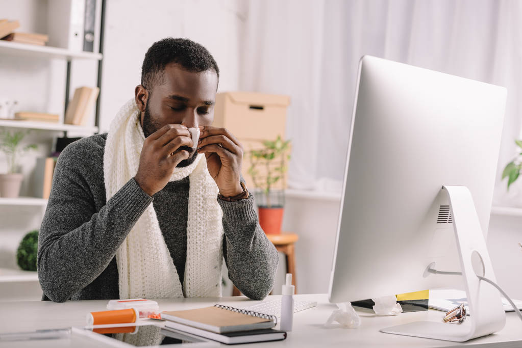 cold african american manager with runny nose holding napkin while sitting at workplace with computer - Photo, Image
