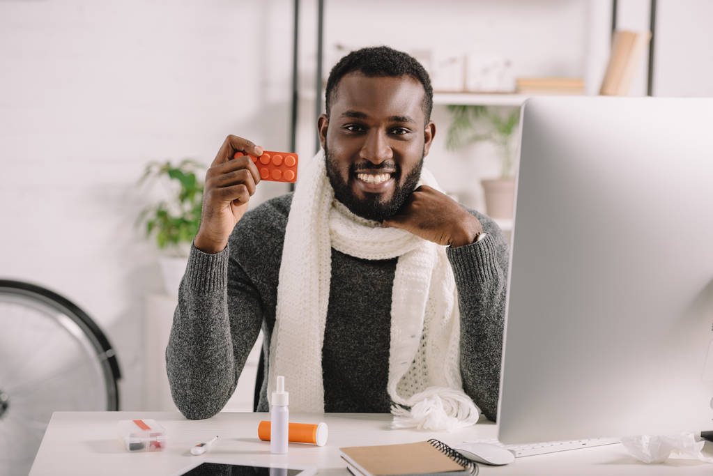 lächelnder kranker afrikanisch-amerikanischer Mann hält Tabletten, während er am Arbeitsplatz mit Medikamenten sitzt - Foto, Bild