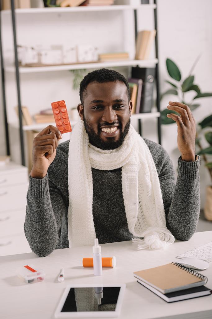 fröhlich kranker afrikanisch-amerikanischer Geschäftsmann hält Tabletten, während er am Arbeitsplatz mit Medikamenten sitzt - Foto, Bild