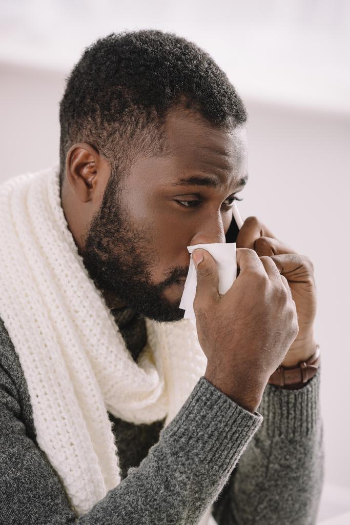 homem americano africano frio em cachecol quente com nariz escorrendo segurando guardanapo enquanto fala no smartphone
 - Foto, Imagem