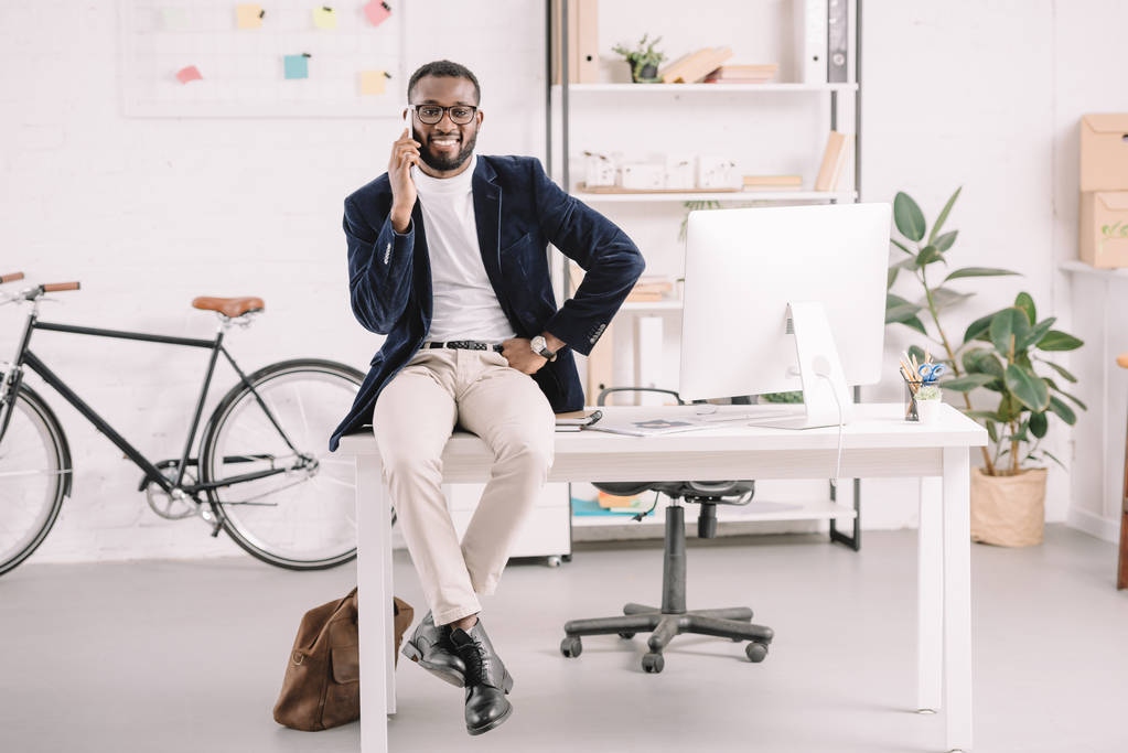 近代的なオフィスにコンピューターのテーブルの上に座ってスマート フォンで話すアフリカ系アメリカ人のビジネスマン - 写真・画像