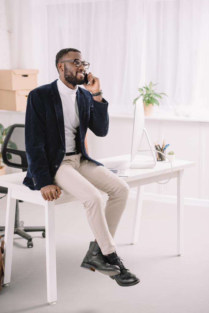 επαγγελματική Αφρικανικός Αμερικανός επιχειρηματίας μιλάει smartphone στο ενώ κάθεται στο τραπέζι στο σύγχρονο γραφείο με υπολογιστή - Φωτογραφία, εικόνα