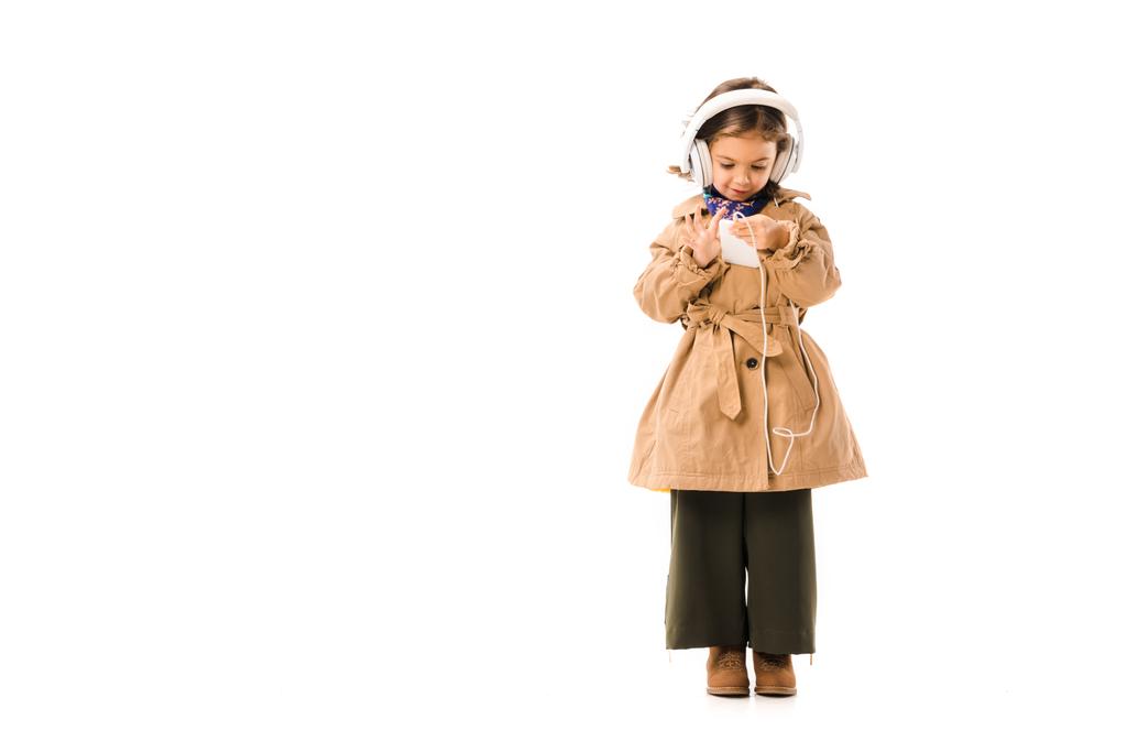 schönes kleines Kind in stylischem Trenchcoat, das mit Kopfhörern Musik hört und das Smartphone isoliert auf weiß verwendet - Foto, Bild