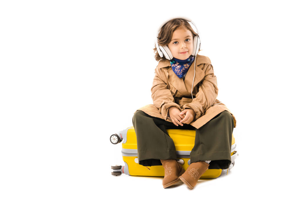 ihana pieni lapsi trench takki kuuntelee musiikkia kuulokkeet ja istuu keltainen matkalaukku eristetty valkoinen
 - Valokuva, kuva