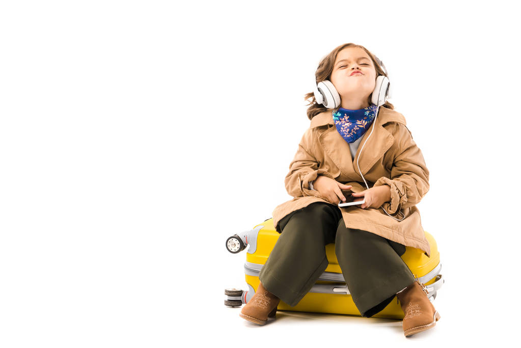 grimacing little child in trench coat listening music with headphones and sitting on yellow suitcase isolated on white - Photo, Image