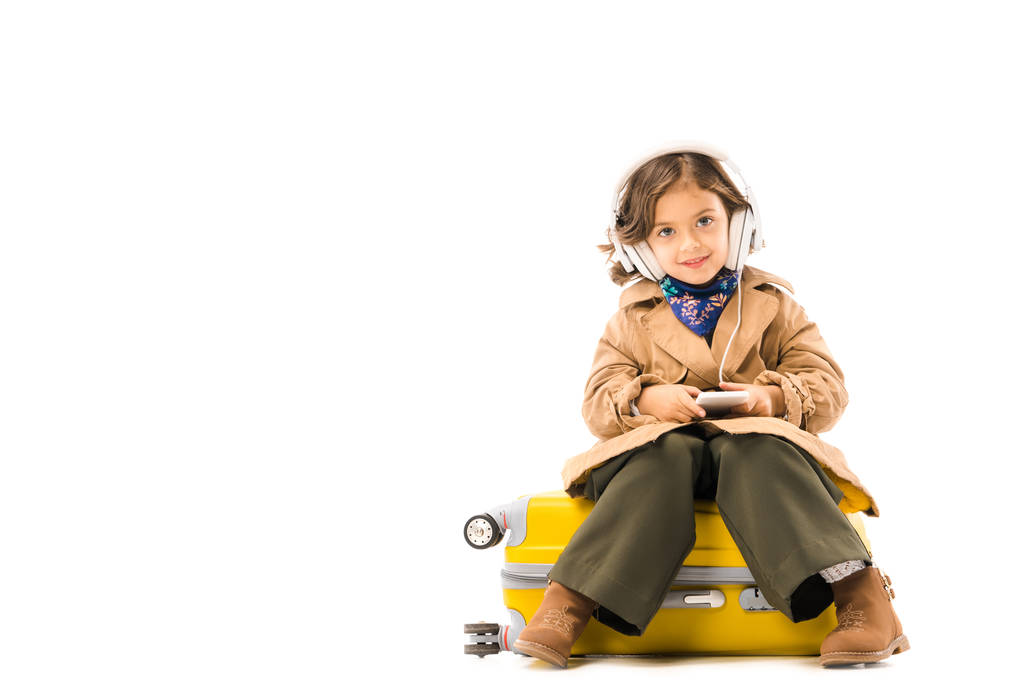 όμορφο μικρό παιδί στην καμπαρντίνα ακούγοντας μουσική με τα ακουστικά και να κάθεται στην βαλίτσα κίτρινο που απομονώνονται σε λευκό - Φωτογραφία, εικόνα