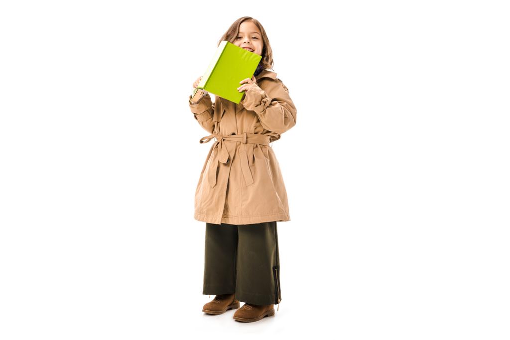 ευτυχισμένο παιδάκι στην καμπαρντίνα με βιβλίο εξετάζει τα φωτογραφικών μηχανών που απομονώνονται σε λευκό - Φωτογραφία, εικόνα