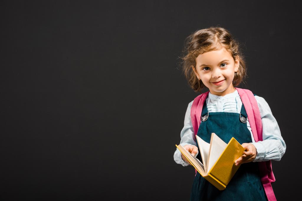 schattig schoolmeisje met rugzak en boek in handen kijken camera geïsoleerd op zwart - Foto, afbeelding