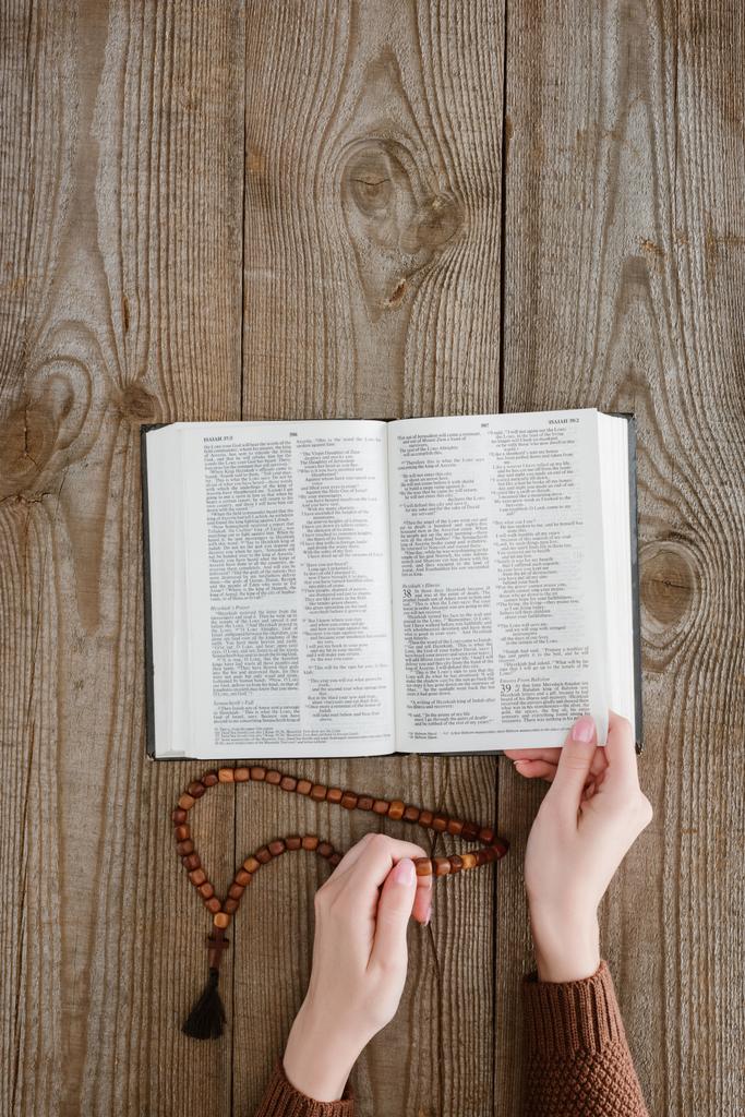 Colpo ritagliato di donna che legge la Sacra Bibbia e tiene perline su un tavolo di legno
 - Foto, immagini