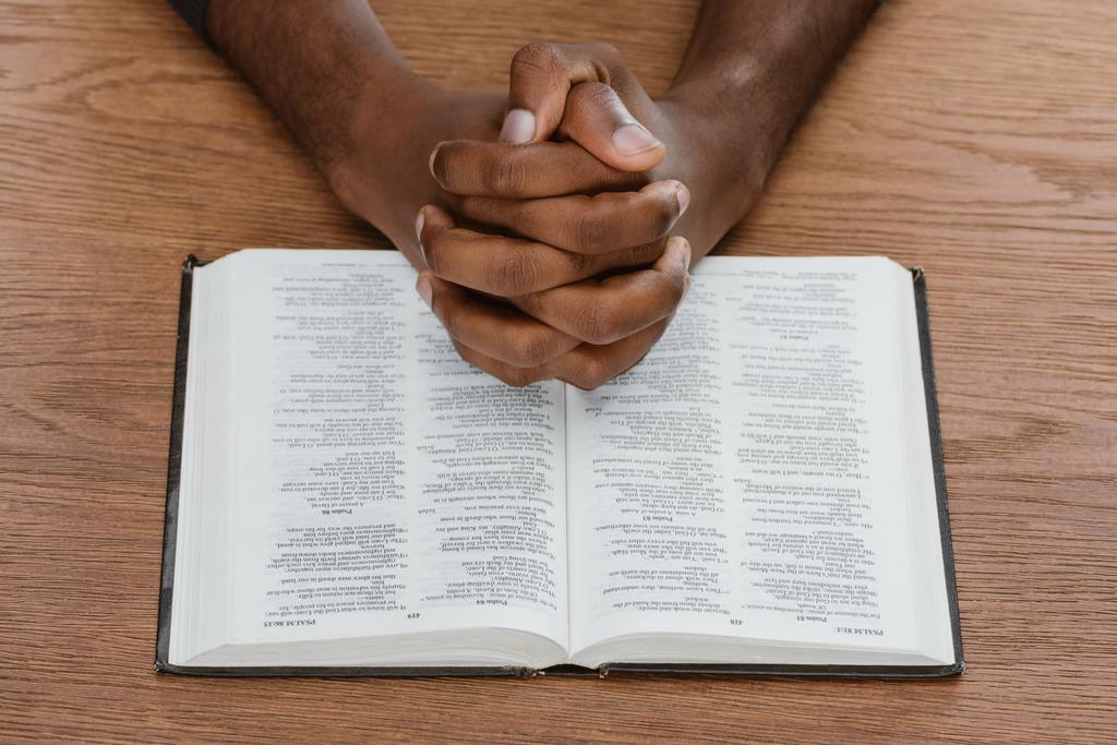 обрізаний знімок афроамериканця, який молиться святою Біблією на дерев'яній поверхні
 - Фото, зображення