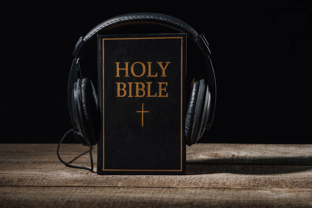 close-up shot of holy bible with headphones standing on wooden table isolated on black - Photo, Image