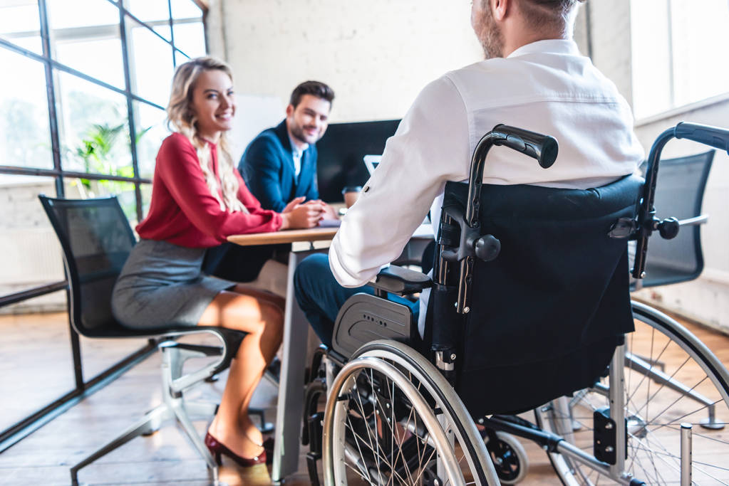 partial view of smiling businesspeople looking at colleague in wheelchair in office - Photo, Image