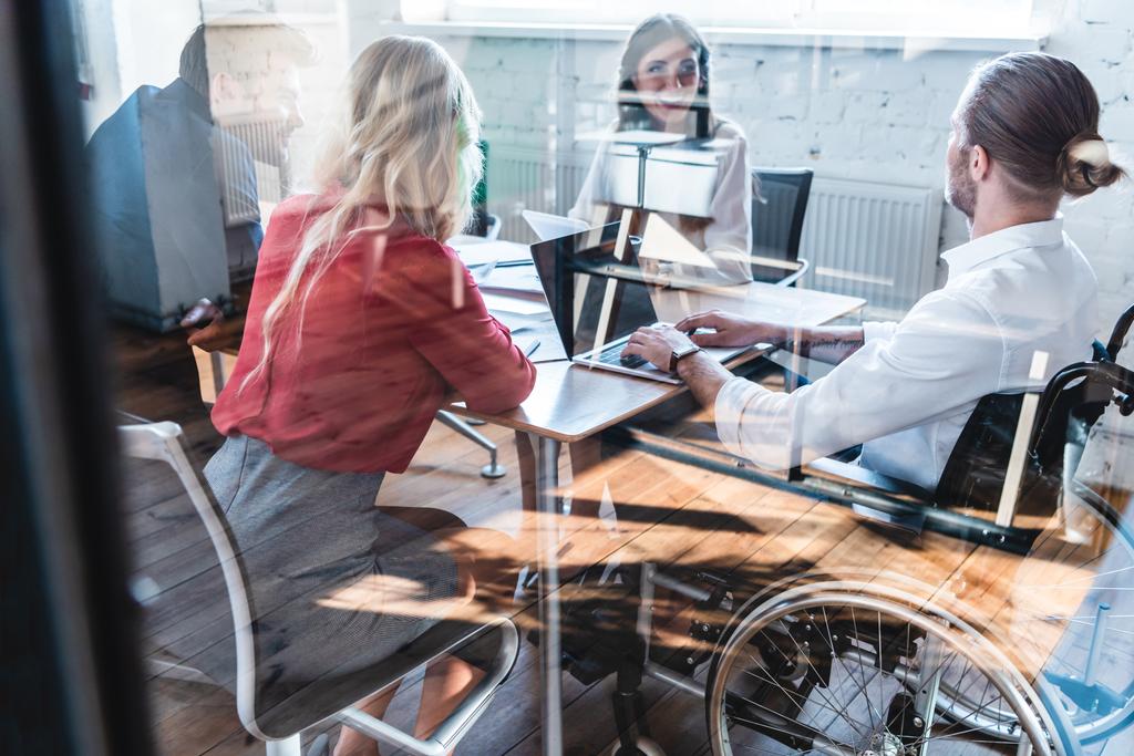 vue à travers le verre de jeune homme d'affaires handicapé en fauteuil roulant travaillant avec des collègues au bureau
  - Photo, image